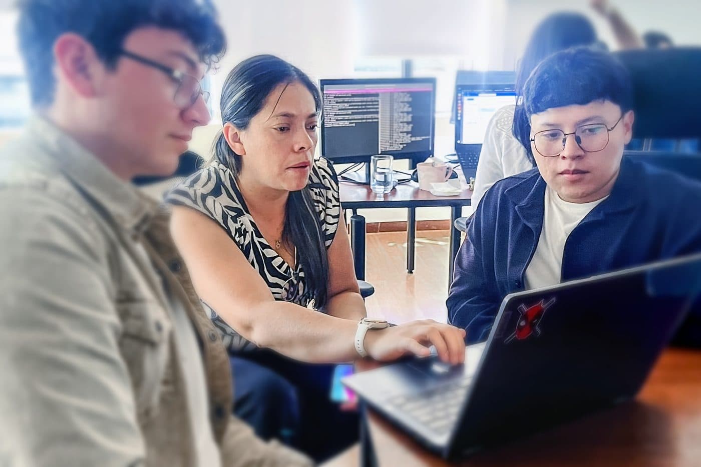 Tres ingenieros de Senirop revisando una compilación juntos frente a una laptop, con una terminal abierta en el monitor detrás, en la oficina de Quito, Ecuador