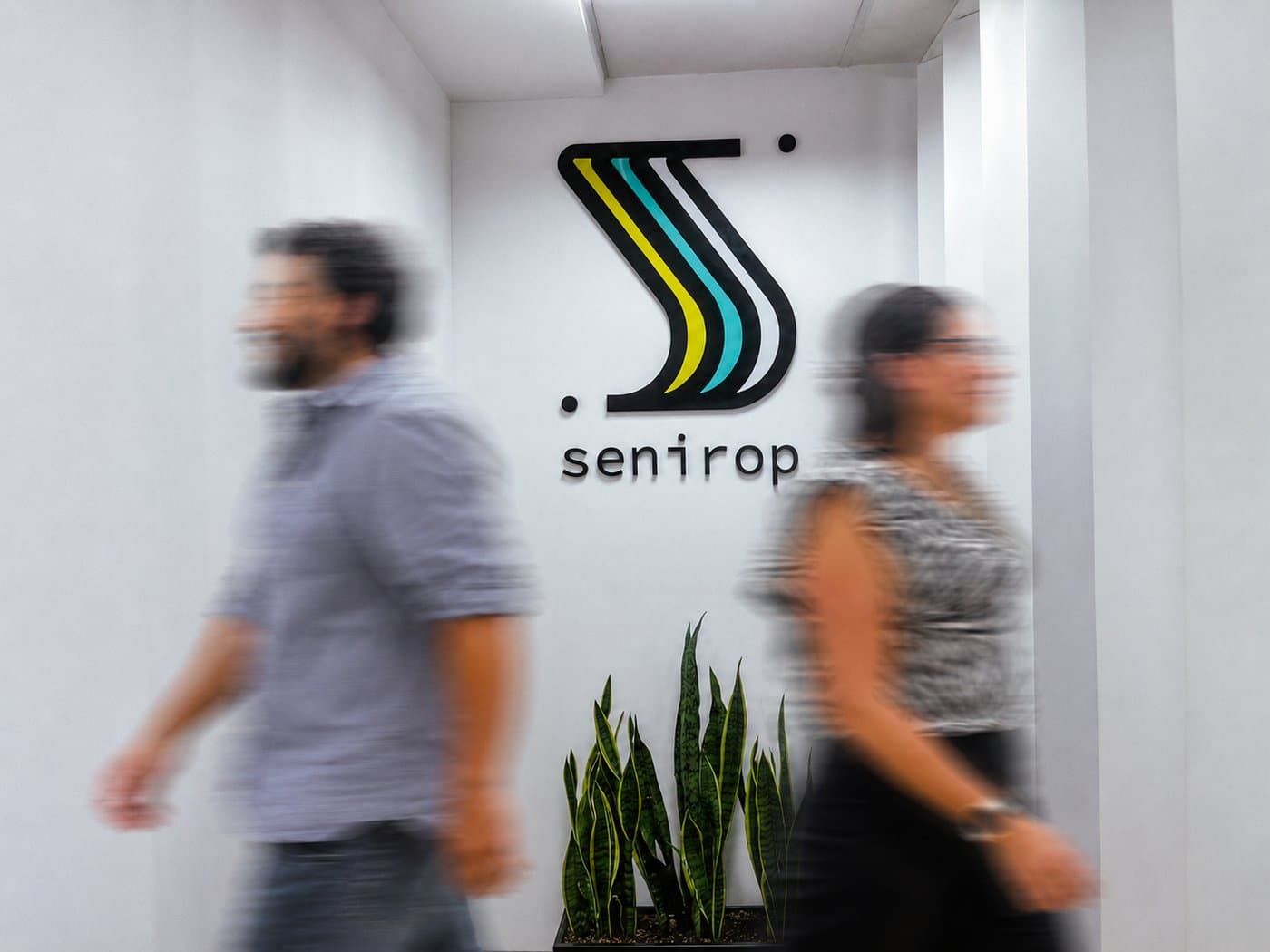Dos miembros del equipo de Senirop caminando frente al muro con el logo de Senirop en la oficina de Quito