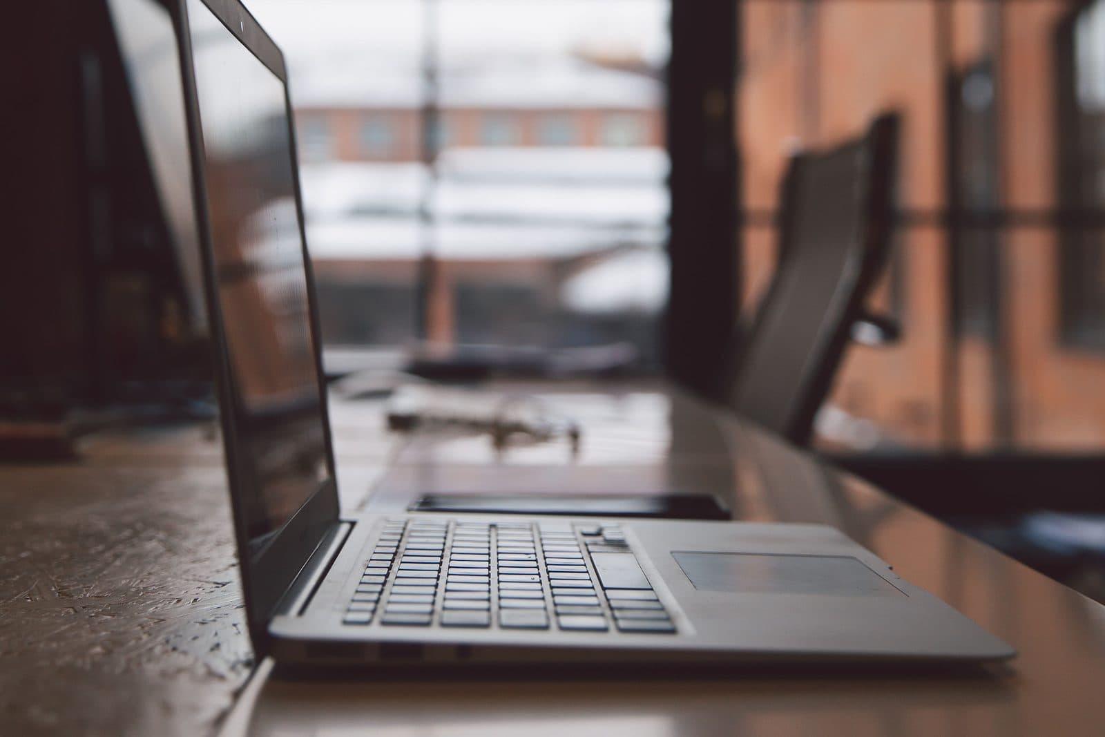 A laptop open on a wooden desk beside a tall factory window, set up for focused technical work