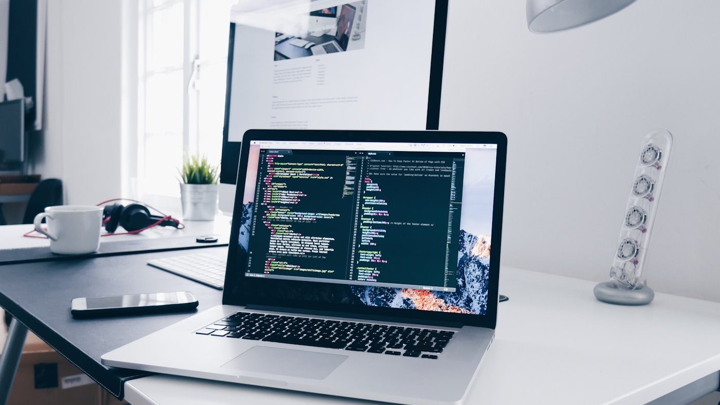 A laptop showing a code editor on a bright desk beside an external monitor and headphones.
