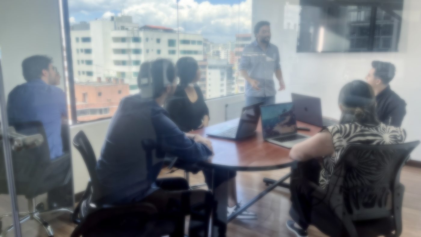 A Senirop consultant leading an AI strategy session for the delivery team around a boardroom table of laptops, with the Quito, Ecuador skyline through the window.